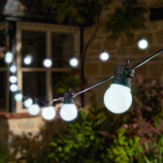 A string of glowing outdoor light bulbs hangs diagonally in the foreground, softly illuminating a garden scene at night, with a blurred house window and greenery in the background.