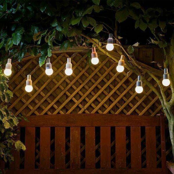 A row of warm white string lights is draped across a wooden lattice fence, glowing softly in a garden at night, with leafy tree branches framing the top of the scene.