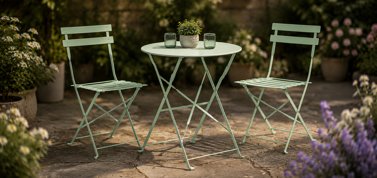 A bright yellow three-piece metal patio bistro set consisting of a round table and two folding chairs, styled on a gray stone patio with green garden foliage in the background.