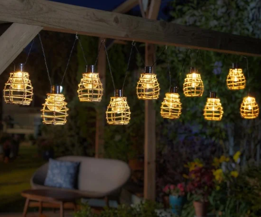 Warm white solar rattan lantern string lights hanging from a wooden pergola, illuminating an outdoor garden seating area at night