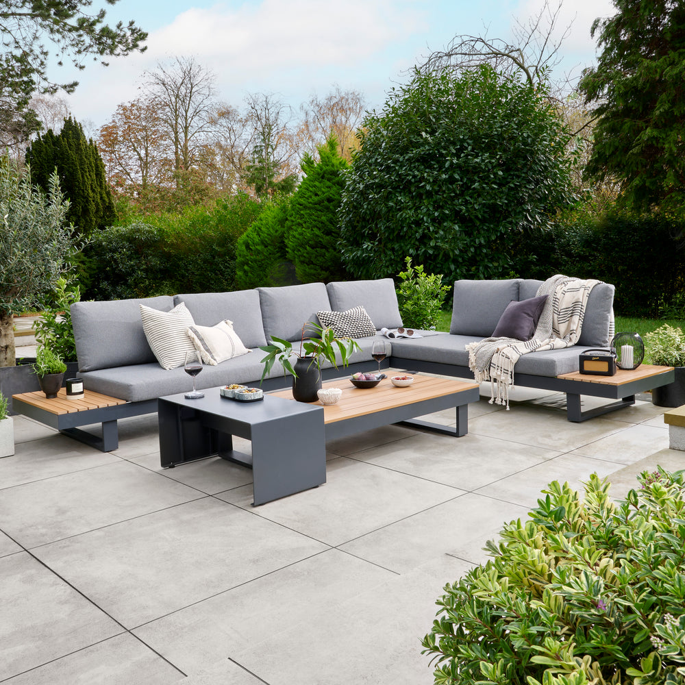 Outdoor patio set with gray sectional sofa, coffee table, and chairs on a paved patio with greenery.