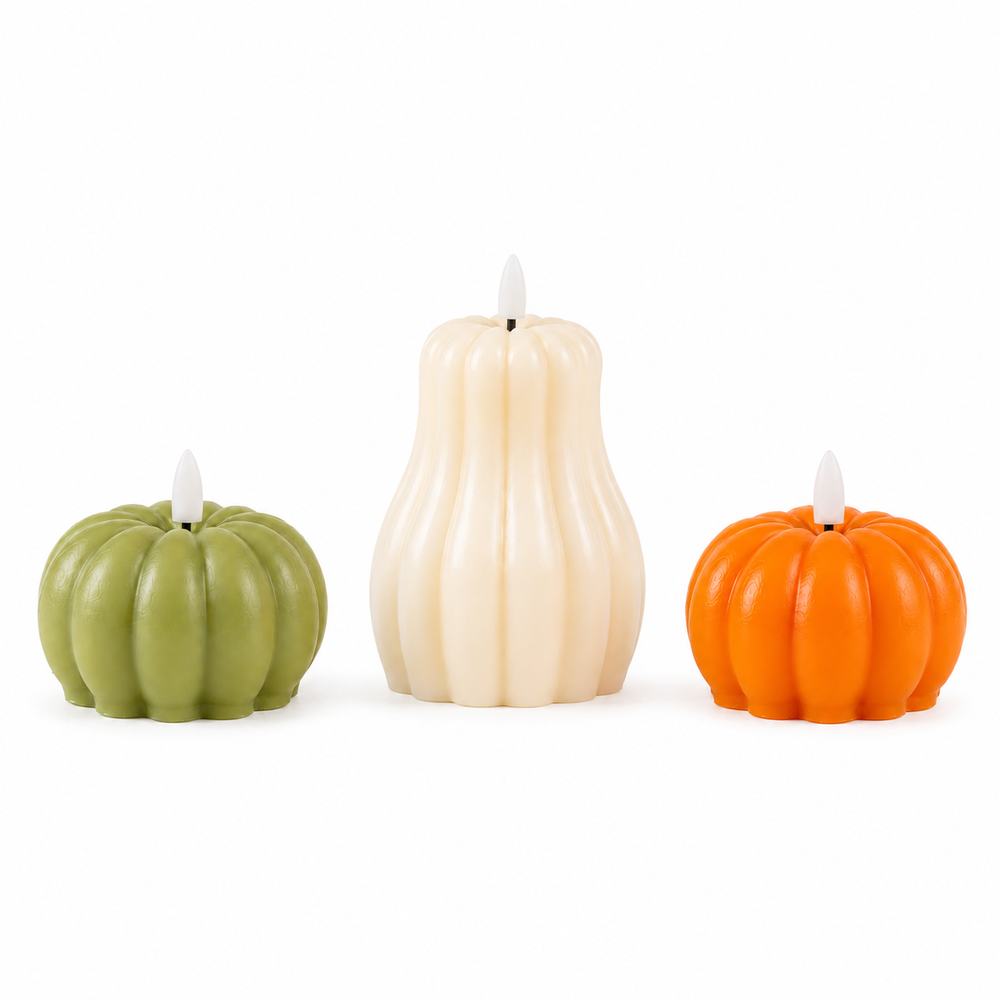 Three flameless LED pumpkin-shaped candles in green, white, and orange are arranged on a burlap table runner with eucalyptus leaves and pinecones in a warm, cozy dining setting.