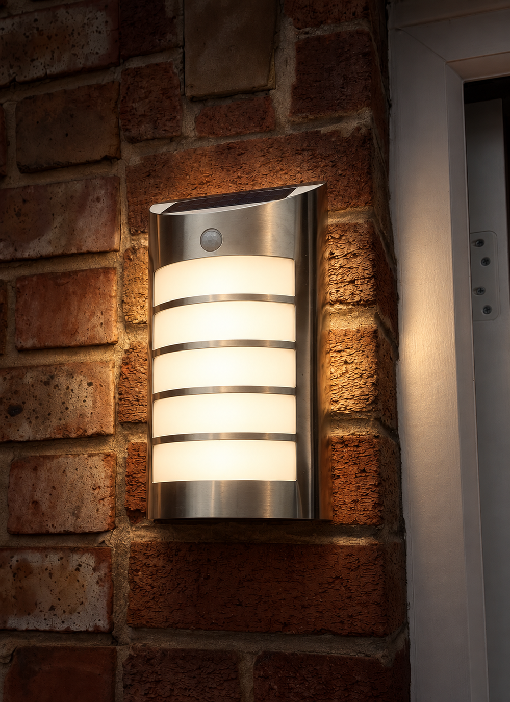 A modern outdoor wall light with a brushed stainless steel finish and horizontal frosted glass panels, mounted on a red brick wall and glowing with warm light.