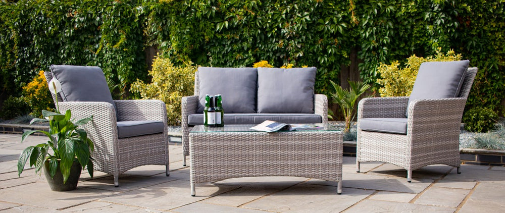 Outdoor patio set with wicker chairs, sofa, and table on a stone patio with greenery in the background.