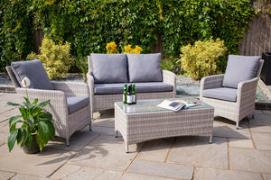 Outdoor patio set with wicker chairs, sofa, and table on a stone patio with greenery in the background.