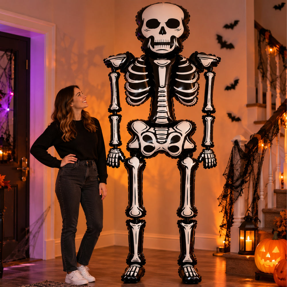 A large Halloween skeleton decoration stands in a decorated home hallway with warm string lights, jack-o'-lantern lanterns, and a small spooky tree.