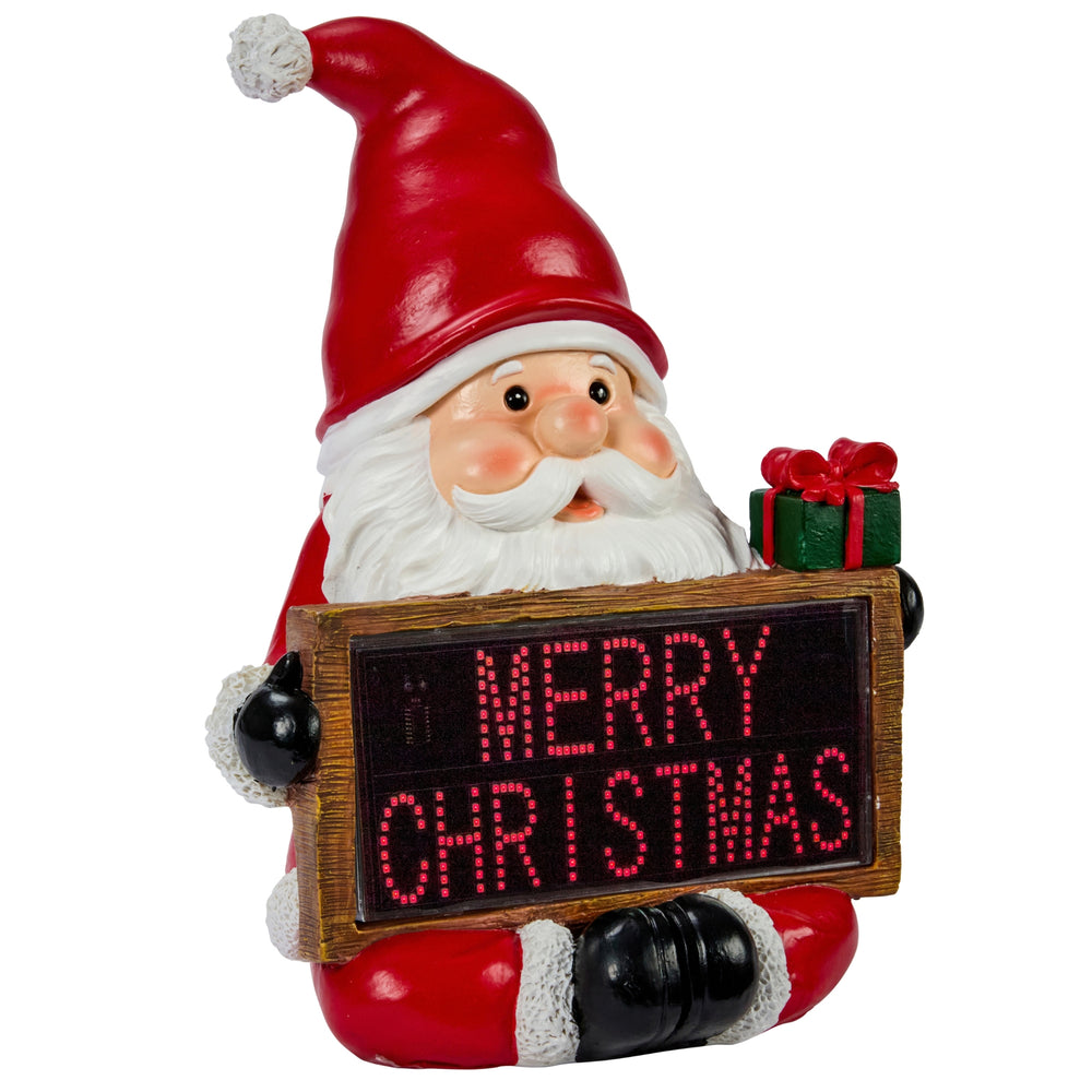 A festive Santa Claus garden statue holding a glowing red LED sign that reads “MERRY CHRISTMAS,” placed outdoors in front of a decorated holiday home with warm lights and a Christmas tree.