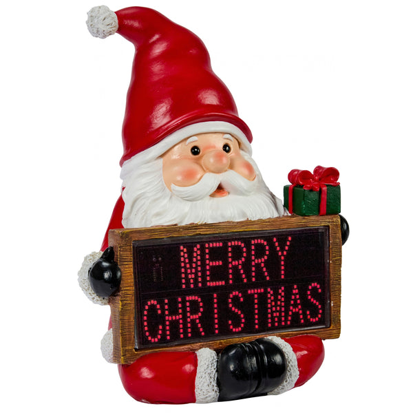 A Santa Claus figurine holding a wooden sign that reads 'MERRY CHRISTMAS' in red LED-style letters, with a small green gift box on his shoulder, shown on a plain white background.