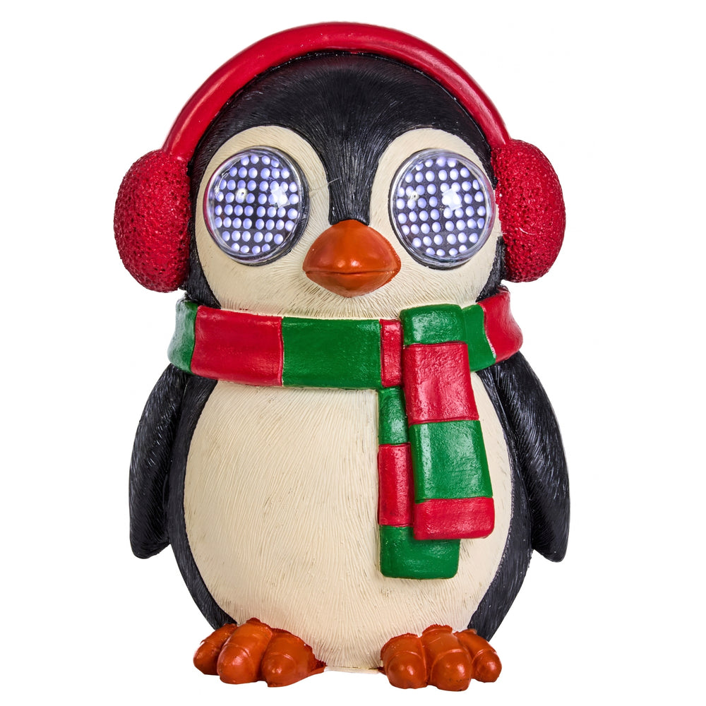 A penguin-shaped decorative figure wearing a red ear warmer hat and a red and green striped scarf, with glowing blue LED-style eyes and orange feet, set in a festive holiday scene with lights and a wrapped gift in the background.