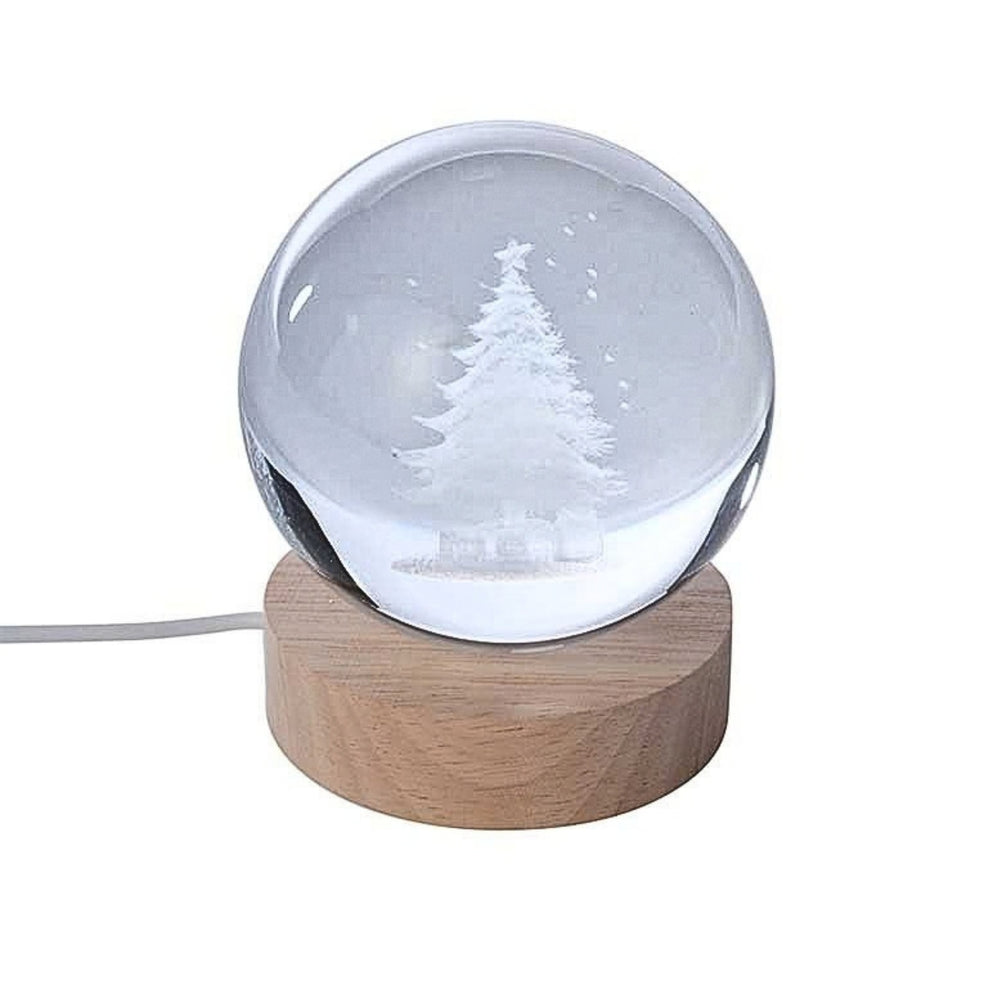 A glowing Christmas tree snow globe lamp with a warm light inside a clear sphere, set on a round wooden base on a tabletop. A blurred holiday plant appears in the background.