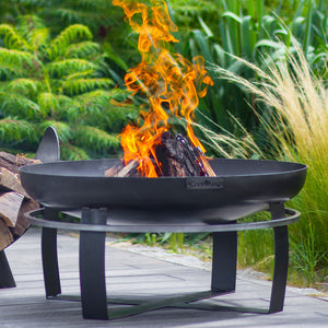 Black fire pit with flames burning on a wooden deck surrounded by greenery