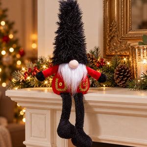 Sitting Christmas guard gonk with a tall black hat, red coat and dangling legs on a mantelpiece beside a festive garland.