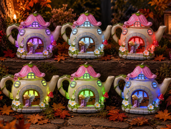 Six fairy teapot garden lights arranged in two rows on a stone surface with autumn leaves. Each teapot-shaped light has a pink roof, a small fairy figure inside, and multicolor LED window lights glowing in different colors including purple, blue, green, and red.