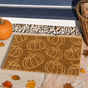A rectangular coir doormat with a repeating pumpkin pattern, placed on a stone patio in a fall-themed setting with a pumpkin, leaves, and a basket nearby.