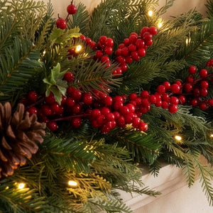 A close-up of a Christmas garland with green pine branches, clusters of red berries, a brown pinecone, and warm white string lights against a soft beige background.