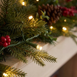 Warm white LED holiday string lights woven through green pine garland with red berries and a pinecone, draped over a white fireplace mantel with a blurred fire in the background.
