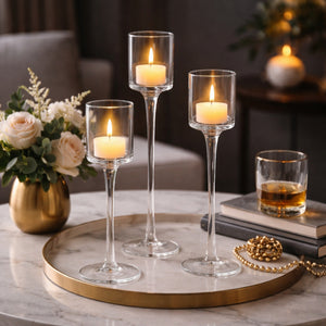 Three clear glass candlestick holders with lit taper candles arranged on a round marble tray with a gold rim, styled in a cozy indoor setting with flowers, books, and a glass of amber liquid.