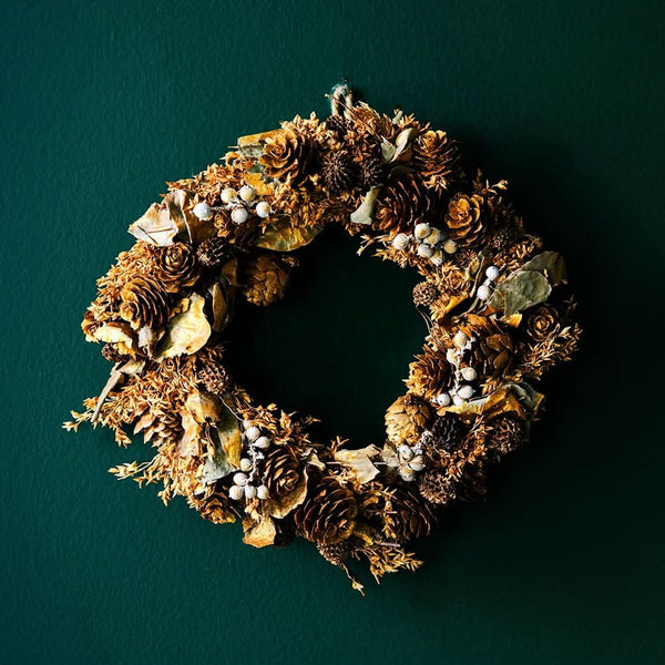 A hanging wreath with a natural orange color, adorned with pine cones and other festive decorations, set against a dark green background.