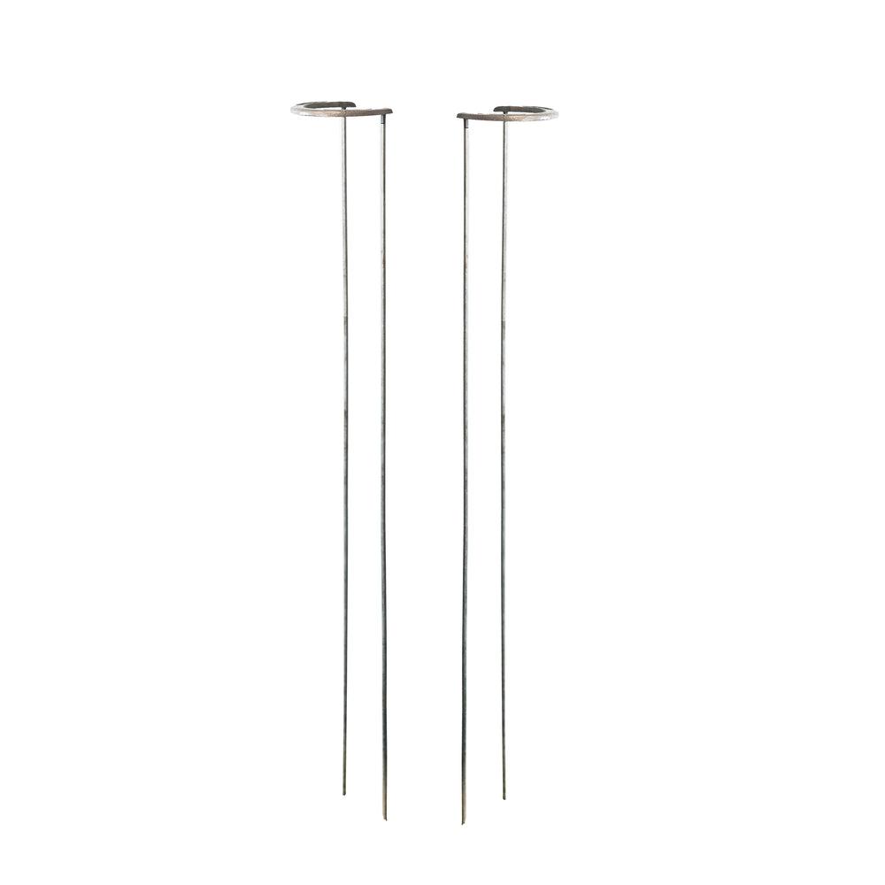Two decorative metal horseshoe garden stakes holding tulip stems, one bright pink tulip and one dark purple tulip, set in a garden with blurred green foliage and soil in the background.