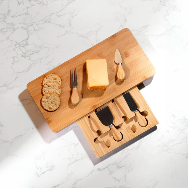 Wooden cheese board set with cheese, crackers, and utensils on a marble surface