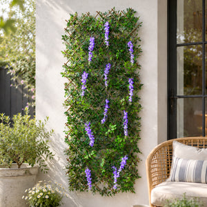 A vertical wall-mounted garden trellis covered in dense green faux ivy with hanging purple wisteria-style flower strands, shown installed on a white exterior wall beside a wicker chair and potted plants.