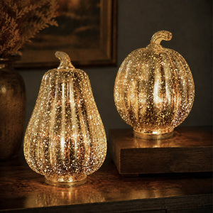 Two decorative mercury glass effect pumpkins with a gold glitter finish, one pear-shaped and one round, lit from within to glow warmly on a dark wooden surface.