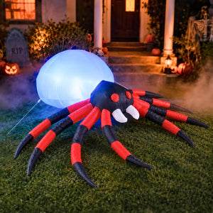 An inflatable Halloween spider decoration with glowing blue eyes and red-and-black striped legs, placed on a lawn in front of a decorated house at night.