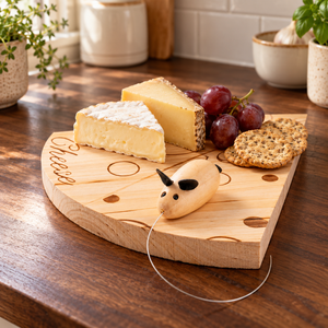 A wooden cheese board with engraved lettering and circular cutouts, styled on a dark wood table with cheese wedges, grapes, crackers, and a small wooden cow cheese pull.