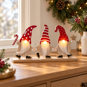 Three wooden Christmas gnomes with glowing white beards and red hats decorated with white hearts and stripes, standing together on a light wood base on a wooden table with warm holiday decor in the background.