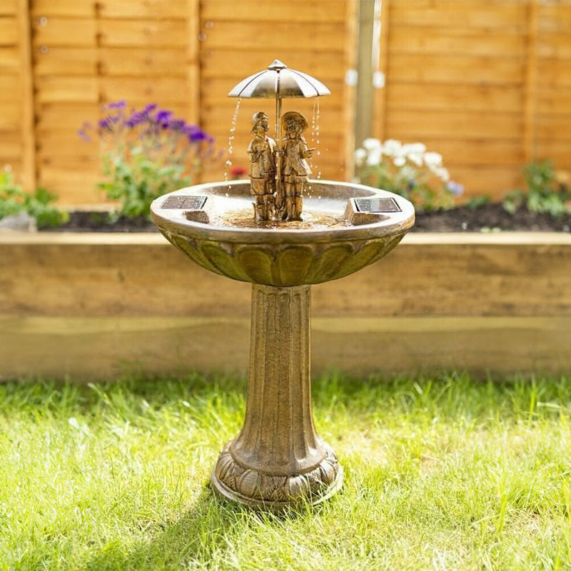 A solar powered water fountain with a bronze finish, featuring a central figure under an umbrella, positioned on a grassy area.