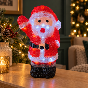 A festive Santa Claus figurine made of red and white textured material, glowing with warm white LED lights, standing on a dark base on a wooden table in a decorated holiday living room.