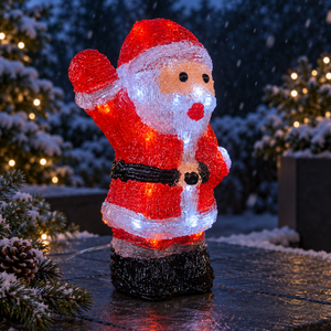 A festive Santa Claus light figurine glowing with warm red and cool white lights, standing on a stone surface in a snowy outdoor setting with blurred Christmas trees in the background.
