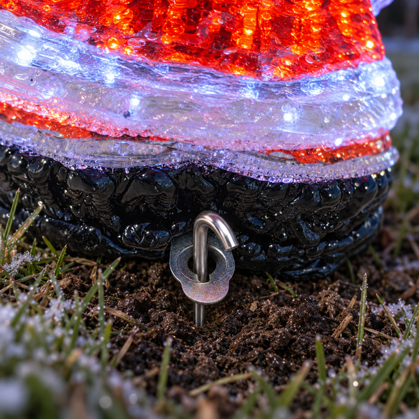 A close-up of a lit Christmas ornament shaped like a boot, with a red and white glowing top, a black textured lower section, and a metal ground spike inserted into dark soil outdoors.