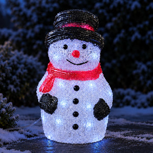 A snowman-shaped outdoor light decoration made of textured white material with glowing bulbs, wearing a black hat with a red band, a red scarf, and black buttons, photographed at night in a snowy setting.