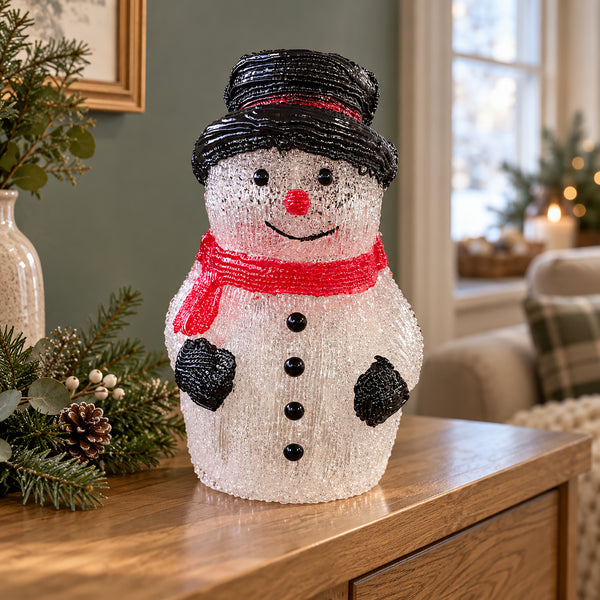A snowman-shaped holiday light decoration with a black hat, red scarf, and black buttons, displayed on a wooden table in a cozy living room setting.