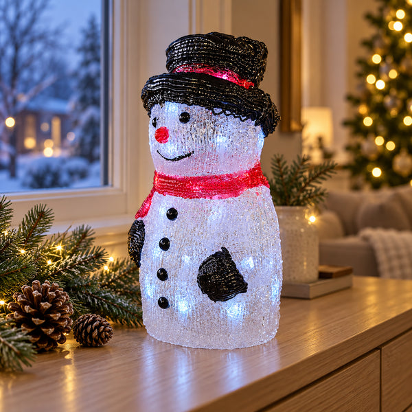 A snowman-shaped LED light decoration with a black hat, red scarf, and glowing white body, displayed on a wooden table in a cozy holiday living room setting.