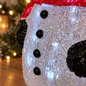 A close-up of a snowman decoration with a white glittery body, black buttons, black ears, and a red glittery hat, photographed indoors with blurred holiday lights in the background.