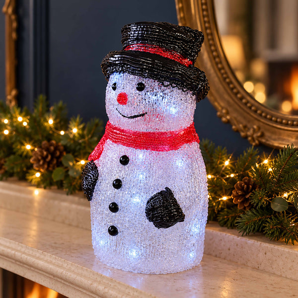 A snowman-shaped holiday light decoration made of black and white sequins, with a red sequin hat band and scarf, standing on a mantel decorated with pine branches and warm lights.
