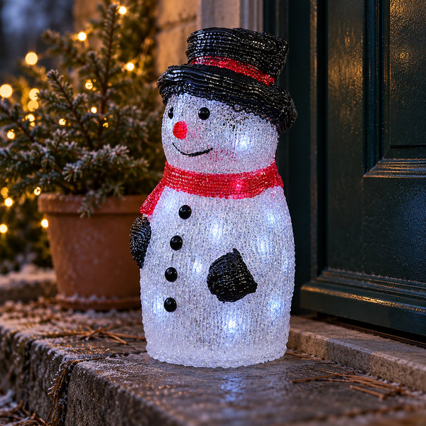 A snowman-shaped outdoor LED light decoration with a black hat, red scarf, and glowing white body, placed on a stone step in front of a dark door with holiday lights in the background.