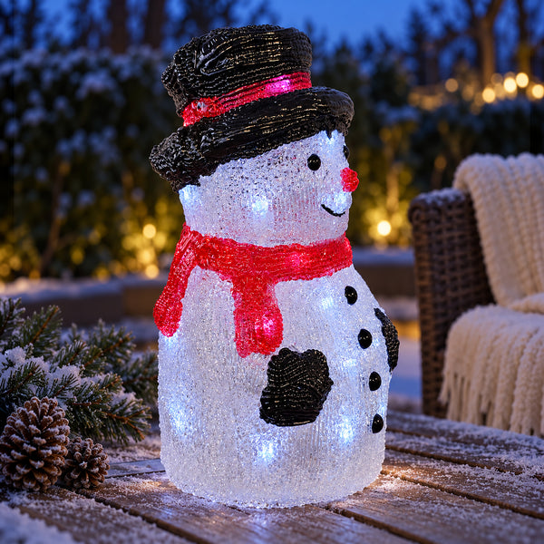 A snowman-shaped outdoor LED light decoration with a black hat, red scarf, and glowing white body, placed on a wooden table in a snowy patio setting at dusk.