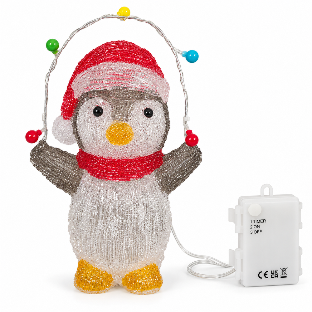 A lighted penguin Christmas decoration standing on a wet stone step in front of a decorated house at night. The penguin is made of glowing white textured material with a red Santa hat, red scarf, yellow beak, and yellow feet, and holds a string of multicolored lights.