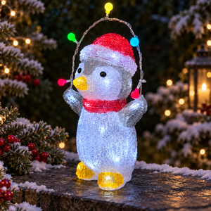A penguin-shaped outdoor light decoration covered in white LED lights, wearing a red Santa hat and red scarf, standing on a snowy surface with multicolored hanging holiday lights in a festive outdoor setting.