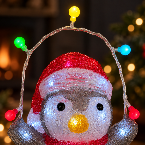 A glittery penguin Christmas decoration wearing a red and white Santa hat, with multicolored LED string lights in red, green, blue, and yellow around it, set against a blurred holiday background.