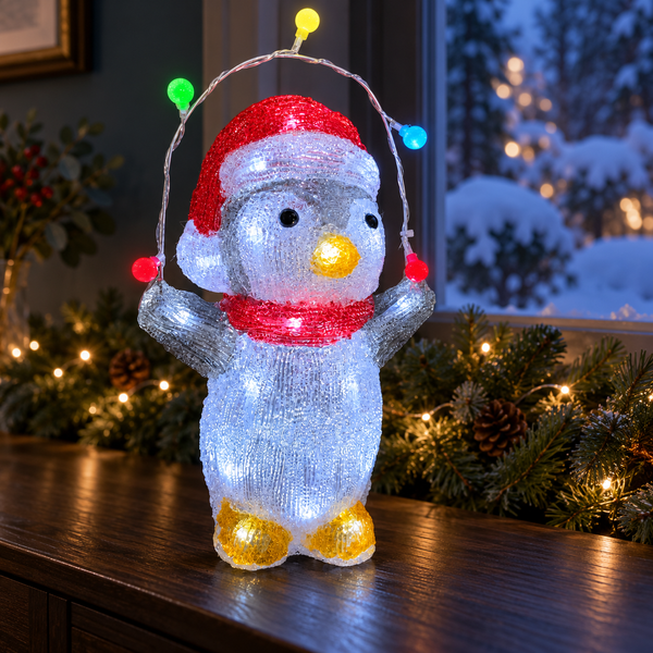 A penguin-shaped LED light decoration stands on a dark wooden surface in a holiday setting. The penguin is white with a red Santa hat, red scarf, yellow beak, and yellow feet, holding a string of multicolored lights above its head.