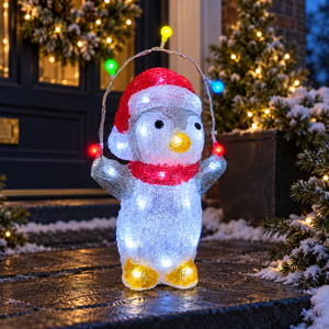 A lighted penguin Christmas decoration standing on a wet stone step in front of a decorated house at night. The penguin is made of glowing white textured material with a red Santa hat, red scarf, yellow beak, and yellow feet, and holds a string of multicolored lights.