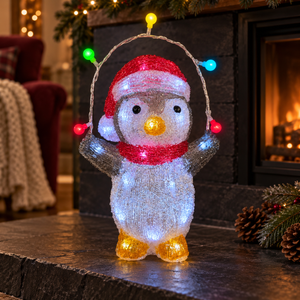 A penguin-shaped holiday light decoration stands on a dark stone surface in front of a fireplace. The figure wears a red Santa hat and red scarf, with multicolored string lights attached above and around it.