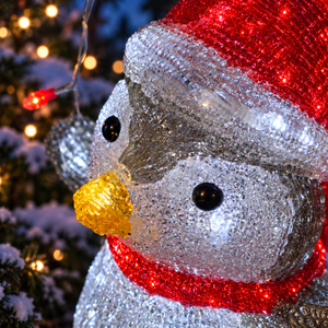 A close-up of a glittery penguin Christmas ornament decorated with a red Santa hat, red scarf, and yellow beak, shown in front of a blurred Christmas tree with warm lights.