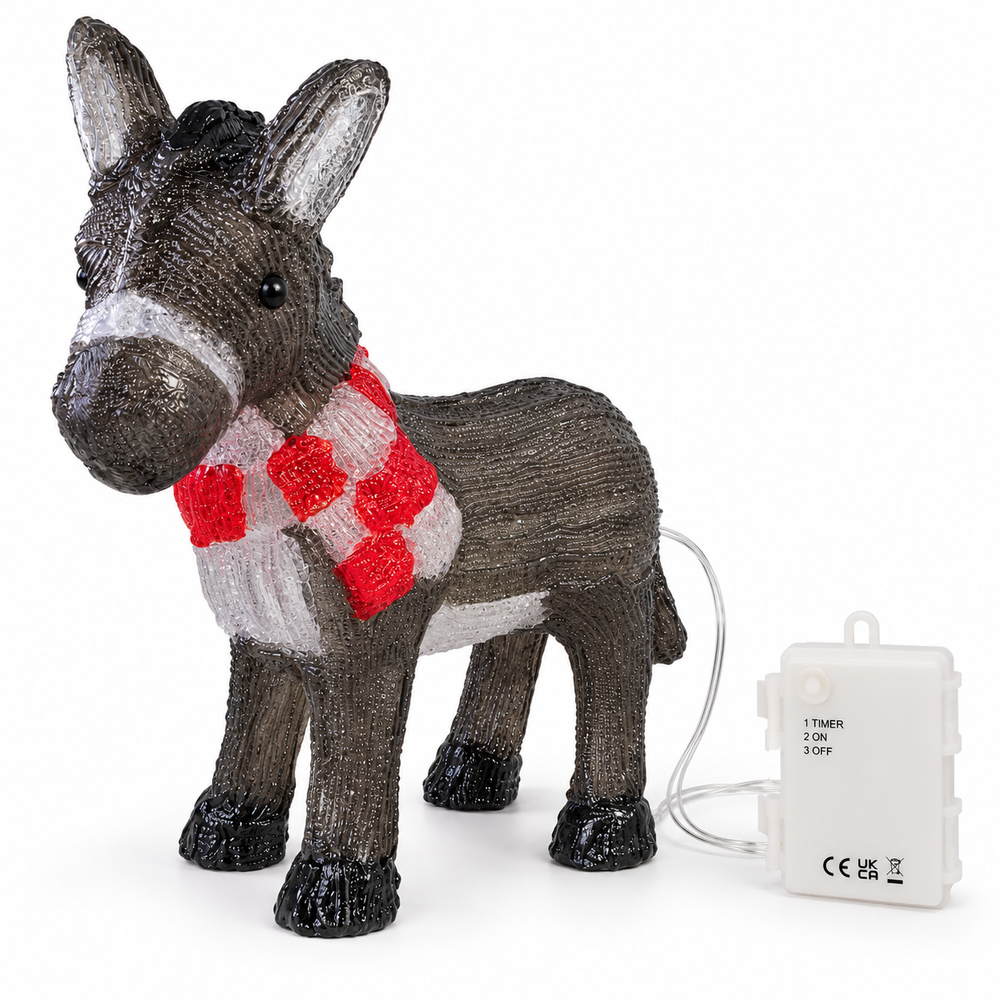A decorative donkey figurine outdoors at night, covered in small white lights and wearing a red-and-white patterned scarf, standing on a stone surface with snowy ground and blurred holiday lights in the background.