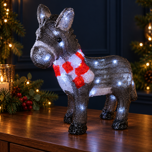A lit donkey-shaped holiday decoration with a red and white striped scarf, standing on a wooden table with Christmas trees and warm lights in the background.