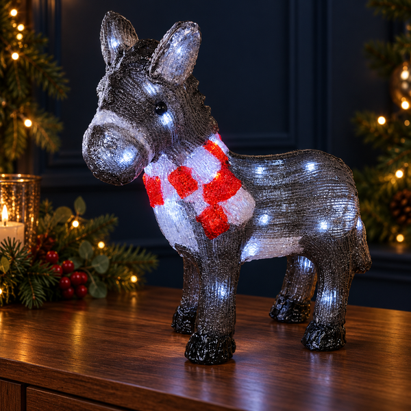 A lit donkey-shaped holiday decoration with a red and white striped scarf, standing on a wooden table with Christmas trees and warm lights in the background.